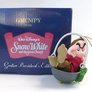 May include: A Grumpy ornament from Walt Disney's Snow White and the Seven Dwarfs. The ornament features Grumpy sitting in a bucket with a grumpy expression on his face. The ornament is part of the Grolier President's Edition.