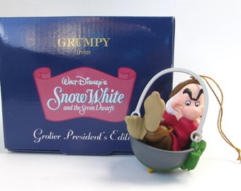 Puede incluir: Un adorno de Grumpy de Blancanieves y los siete enanitos de Walt Disney. El adorno presenta a Grumpy sentado en un cubo con una expresión de enfado en su rostro. El adorno forma parte de la edición del presidente Grolier.