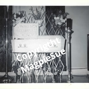May include: A white casket with floral arrangements on either side, a silver candelabra, and a television in the background.