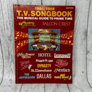 May include: A 1984/1985 T.V. Songbook titled The Musical Guide to Prime Time. The red cover features a central illustration of a television set shaped like a piano with musical notes, surrounded by logos for shows like Cheers, Falcon Crest, Hill Street Blues, Hotel, Entertainment Tonight, Paper Dolls, Maggie Briggs, Dynasty, Fame, St. Elsewhere, Newhart, The Jeffersons, and Dallas.