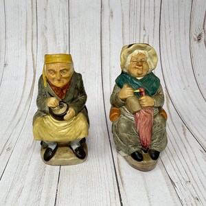 Peut inclure: Deux figurines en céramique de personnes âgées. La figurine de gauche porte un chapeau jaune et une veste verte. La figurine de droite porte un chapeau blanc et une veste verte. Les deux figurines tiennent des objets dans leurs mains.