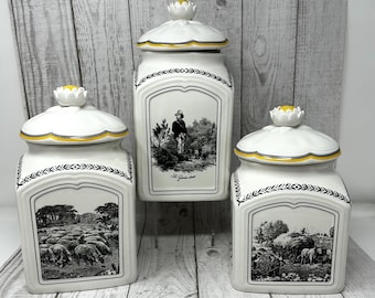 May include: Three white ceramic canisters with lids featuring yellow and black trim. Each canister displays a black and white pastoral scene. The central canister is taller and features the text Le Garde-bois, while the others show rural farm life.
