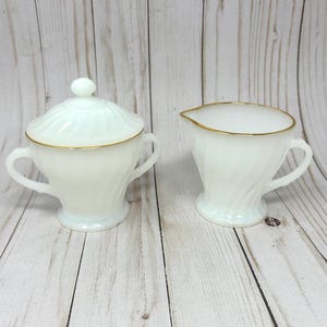 May include: A white milk glass sugar bowl with a matching lid and a creamer jug. Both pieces feature a swirling ribbed pattern, two handles on the sugar bowl, one handle on the creamer, and a thin gold trim along the top edges.