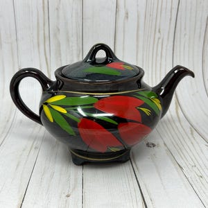 May include: A black ceramic teapot with a matching lid. The teapot is decorated with red, yellow, and green floral designs. It has a curved handle and a spout. The teapot sits on a wooden surface.