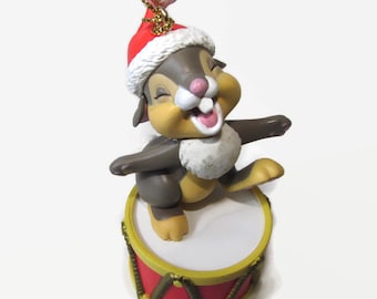 Puede incluir: Un adorno navideño con un conejo marrón y blanco que lleva un gorro de Papá Noel rojo y toca un tambor rojo y dorado.