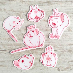 Puede incluir: Seis pegatinas blancas con ilustraciones de ratas en línea roja en varias poses. Una rata lleva una corona de corazón. Las pegatinas están sobre una superficie de madera clara.