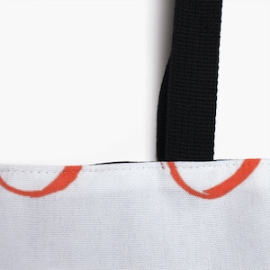 May include: Close-up of a white canvas tote bag with black straps. The bag has a red and white abstract design.