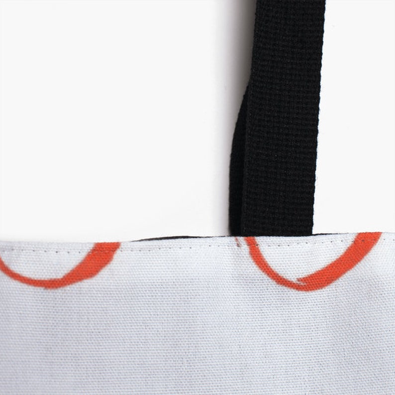 May include: Close-up of a white canvas tote bag with black straps. The bag has a red and white abstract design.