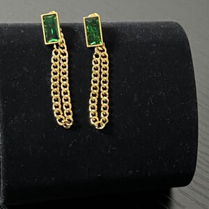 Puede incluir: Un par de pendientes dorados con piedras verdes rectangulares y detalles de cadena colgante. Los pendientes se exhiben sobre una superficie de terciopelo negro, mostrando su dise&ntilde;o y contraste de color.