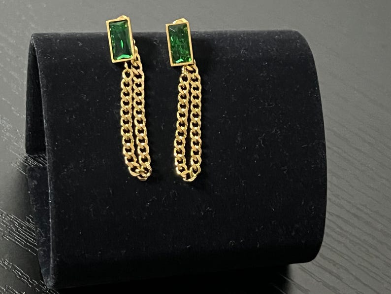 Puede incluir: Un par de pendientes dorados con piedras verdes rectangulares y detalles de cadena colgante. Los pendientes se exhiben sobre una superficie de terciopelo negro, mostrando su dise&ntilde;o y contraste de color.