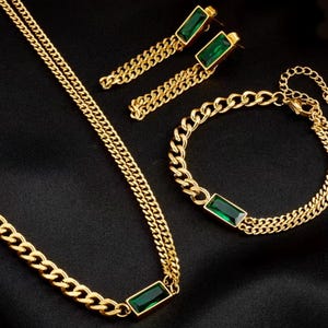 Puede incluir: Un conjunto de joyas doradas que incluye collar, pulsera y pendientes. Cada pieza tiene un dise&ntilde;o de cadena y una piedra rectangular de color verde esmeralda. El conjunto se muestra sobre una superficie negra.