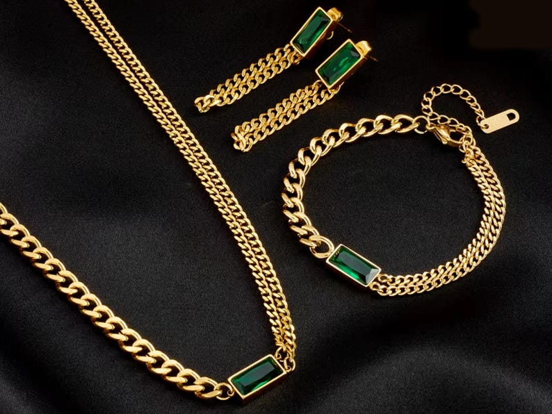 Puede incluir: Un conjunto de joyas doradas que incluye collar, pulsera y pendientes. Cada pieza tiene un dise&ntilde;o de cadena y una piedra rectangular de color verde esmeralda. El conjunto se muestra sobre una superficie negra.