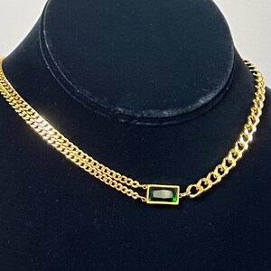 Puede incluir: Collar dorado con un colgante de piedra preciosa verde talla esmeralda. El collar presenta un dise&ntilde;o de doble cadena en un lado y una cadena de eslabones en el otro. La joya se exhibe sobre un maniqu&iacute; negro.