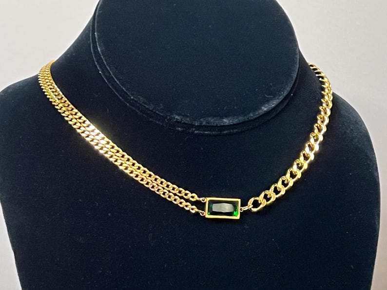 Puede incluir: Collar dorado con un colgante de piedra preciosa verde talla esmeralda. El collar presenta un dise&ntilde;o de doble cadena en un lado y una cadena de eslabones en el otro. La joya se exhibe sobre un maniqu&iacute; negro.