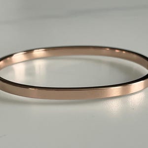 May include: Rose gold bangle bracelet. This simple, elegant bracelet is a solid band of rose gold metal. The bracelet is a complete circle and has a smooth, polished finish. The bangle is a classic jewelry piece.