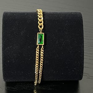 Puede incluir: Una pulsera dorada con un colgante rectangular de piedra preciosa verde esmeralda. La pulsera presenta un dise&ntilde;o de cadena con un colgante central y cadenas colgantes. La joya se exhibe sobre un fondo negro.