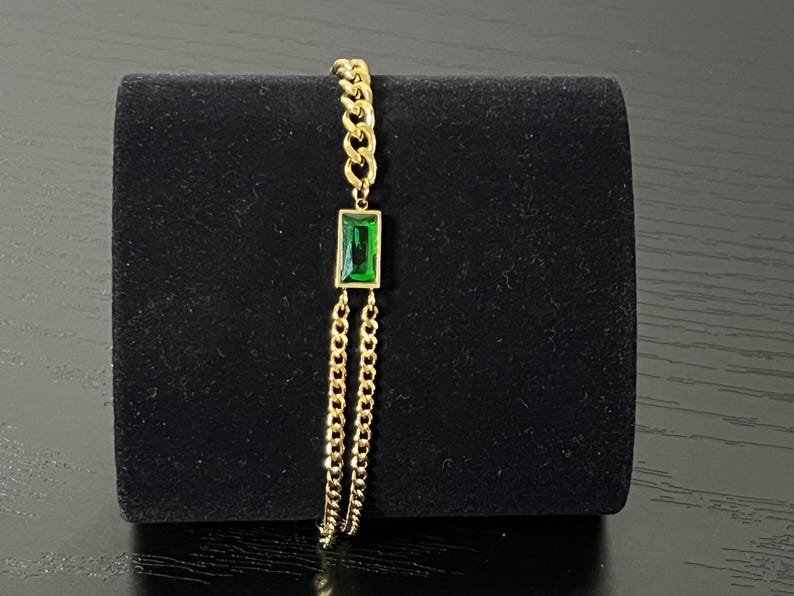 Puede incluir: Una pulsera dorada con un colgante rectangular de piedra preciosa verde esmeralda. La pulsera presenta un dise&ntilde;o de cadena con un colgante central y cadenas colgantes. La joya se exhibe sobre un fondo negro.