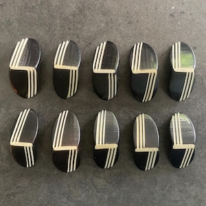 Op de afbeelding: Tien zwarte en crèmekleurige ovale decoratieve objecten. Elk object is verdeeld in twee secties, met een crèmekleurige sectie en een zwarte sectie. Drie crèmekleurige strepen staan op de zwarte sectie.