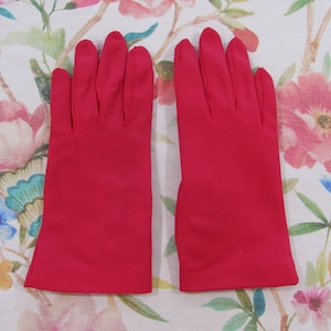 May include: A pair of bright red gloves. The gloves are made of a smooth, solid material and are displayed on a floral patterned background. The gloves are a classic style, with a fitted design and a slightly flared cuff.