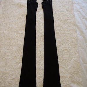 Puede incluir: Un par de guantes largos negros, probablemente para ocasiones formales, se encuentran sobre una superficie blanca estampada. Los guantes se extienden más allá de la muñeca, cubriendo una parte significativa del brazo. La tela es de color sólido.