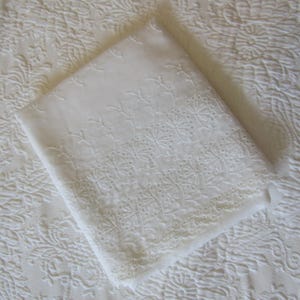 以下が含まれることがあります： 縁に複雑なアイレット刺繍が施された、折りたたまれた白い布の正方形。このテキスタイルは、小さな円形の切り抜きと細かいステッチが特徴の繊細な花柄で、全体的に清潔で均一な白色を示しています。