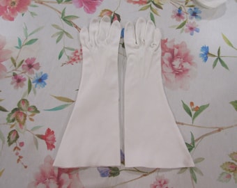 May include: A pair of long, white gloves. The gloves are made of a smooth material, possibly satin, and extend past the wrist. The fingers are finely detailed, and the gloves are displayed on a floral patterned surface.
