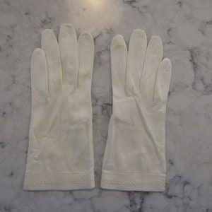 May include: A pair of off-white gloves, likely leather, are laid flat against a marble surface. The gloves have a classic design with a stitched cuff and individual finger slots. The gloves are clean and appear to be new.