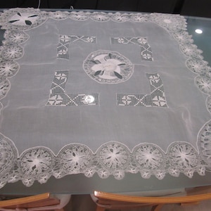 May include: A square white lace tablecloth featuring intricate needlework, a central circular floral embroidery motif, geometric corner patterns, and a wide scalloped border with detailed circular lace medallions along all four edges.