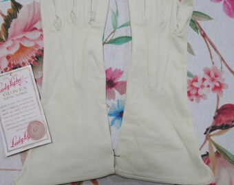 May include: A pair of off-white gloves, likely vintage, with a tag from "Lady Lyke's Gloves New York." The gloves are made of a smooth material and have a classic design. The gloves are laid out on a floral patterned fabric.