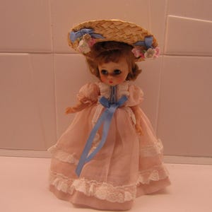 May include: A doll with brown hair wearing a light pink dress with white lace trim and a blue ribbon sash. The doll wears a straw hat decorated with blue ribbons and small pink and white flowers. The doll has painted facial features.