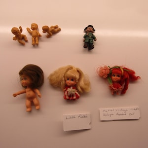 Könnte beinhalten: Eine Sammlung von sechs kleinen Puppen, darunter drei beige Kunststoff-Miniaturfiguren, eine Puppe in grüner Uniform, eine Puppe mit braunem Haar, eine blonde Puppe in rot-weißem Kleid und eine rothaarige Puppe mit rosa Blumenaccessoire und Etiketten.