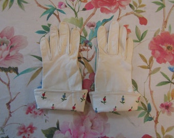 Puede incluir: Un par de guantes color crema con puño bordado floral. Los guantes están hechos de un material suave y presentan un delicado diseño de flores rojas, azules y rosas. Los guantes son probablemente para ocasiones formales.