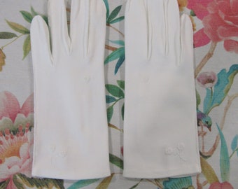 Puede incluir: Un par de guantes blancos impecables con delicados bordados florales cerca de la muñeca. Los guantes están colocados sobre una superficie con estampado floral, mostrando su textura suave y diseño clásico. Un accesorio atemporal.