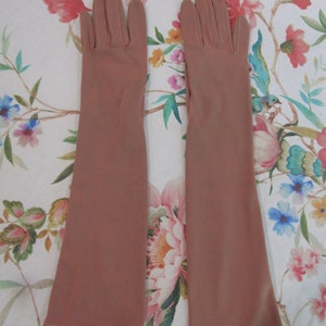 Puede incluir: Un par de guantes largos color rubor. Los guantes están hechos de una tela suave y se extienden más allá de la muñeca. Los guantes están colocados sobre una superficie con estampado floral. Los guantes son de un tono neutro apagado.