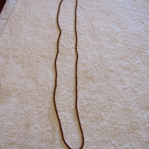 May include: A long, brown beaded necklace. The necklace is made of small, round beads and is lying on a white, patterned surface. The beads have a slightly iridescent quality, catching the light.