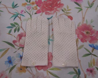 May include: A pair of off-white, crocheted gloves with a button detail at the wrist. The gloves have a delicate, open weave pattern. The background features a floral fabric with pink and red flowers.