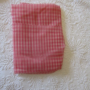 May include: A folded rectangular fabric piece featuring a pink and white gingham check pattern. The textile has a semi-sheer, lightweight appearance with a consistent grid design throughout the surface, showing clear horizontal and vertical lines.