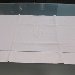 May include: A rectangular white table runner featuring a central embroidered monogram with the letters "JS" enclosed in a diamond shape. The fabric has a horizontal hemstitch detail running across the lower section, creating a decorative border.