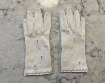 Puede incluir: Un par de guantes blancos con delicado bordado floral rosa. Los guantes presentan pequeñas flores bordadas en la tela y un borde floral detallado en la muñeca. Los guantes están hechos de un material suave y liso.