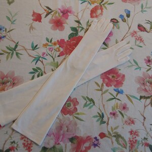 Peut inclure: Une paire de longs gants blancs, en tissu doux et extensible. Les gants sont posés à plat sur une surface à motifs floraux.