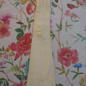 May include: A pair of long, cream-colored gloves. The gloves are made of a soft material and extend well past the wrist. The gloves are lying on a floral patterned surface with pink, red, yellow, and blue flowers.