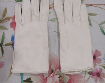 May include: A pair of off-white leather gloves. The gloves are laid flat, showing the front and back. The gloves have a classic design with individual finger slots and a stitched hem. The background is a floral pattern.