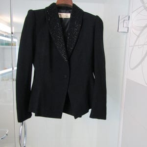Puede incluir: Un blazer negro con cuello con cuentas, colgado de una percha de madera. El blazer tiene un cierre de un solo botón y hombros acolchados. El cuello está adornado con pequeñas cuentas oscuras. La prenda se muestra sobre un fondo blanco.