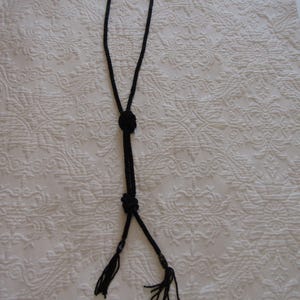 May include: A black beaded necklace with a knotted design. The necklace features a loop, a central knot, and two tassels at the bottom. The tassels have small beads at the end. The necklace is displayed on a white, patterned surface.