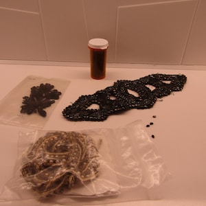 May include: An assortment of craft supplies including a black beaded mask, a brown pill bottle, and various embellishments in clear plastic bags. The items are arranged on a white surface.