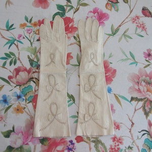 Op de afbeelding: Een paar crèmekleurige, lange leren handschoenen. Elke handschoen heeft een geborduurd strikdesign op de onderarm en een decoratieve lus bij de pols. De handschoenen worden gepresenteerd op een bloemmotief achtergrond.