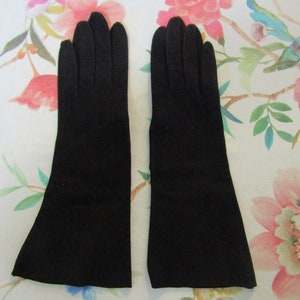 May include: A pair of long, black gloves. The gloves are made of a smooth material and have a classic design. The gloves are displayed on a floral patterned surface with pink and red flowers.