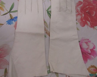 May include: A pair of off-white leather gloves. The gloves are long, extending past the wrist, and have a classic design. The leather appears smooth and the gloves are laid flat, showcasing their shape and construction. The gloves are on a floral background.