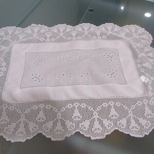 May include: A rectangular white placemat featuring a central fabric panel with delicate white floral embroidery and cutwork details, surrounded by a wide, intricate white crocheted lace border with a repeating bell-shaped pattern.