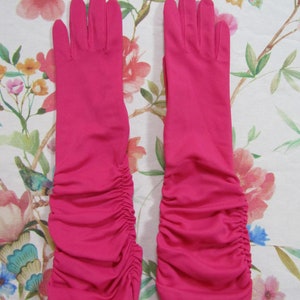 May include: A pair of bright pink, long opera gloves. The gloves are made of a smooth fabric and feature a ruched design on the lower arm. The gloves are displayed on a floral patterned background.